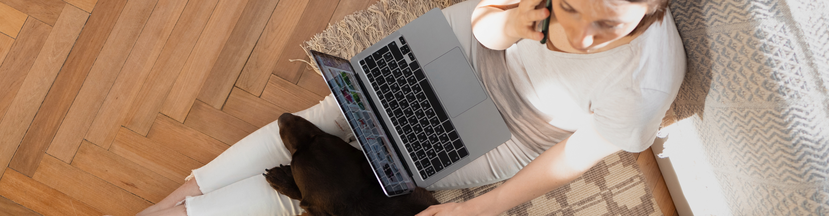 Woman working on a laptop while on the phone, sitting on a cozy rug with a dog, highlighting the warmth and assurance of the Beautiful Guarantee for flooring.